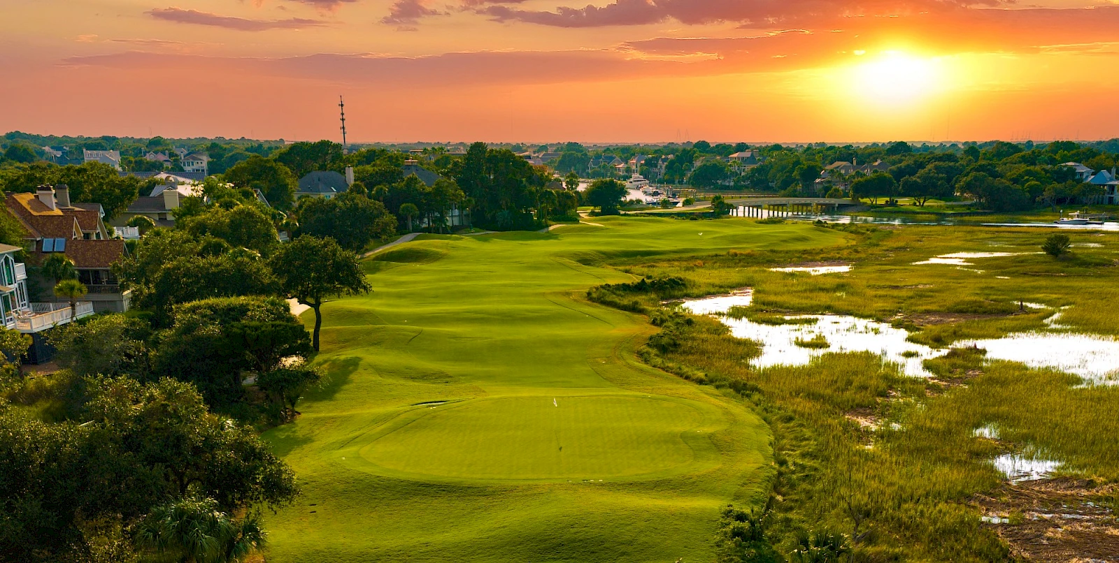 A sunlit golf course with a green fairway, surrounding trees, water-filled marshes, and a calm neighborhood in the background at sunset.