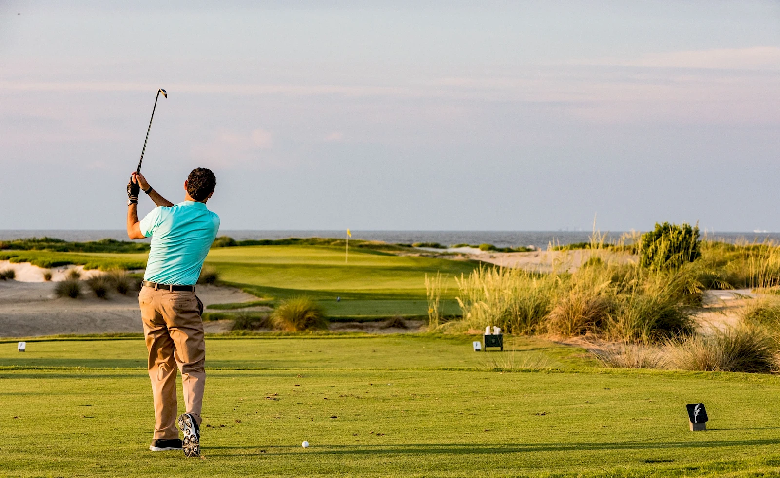 A golfer in a blue shirt is swinging a club on a sunny tee box, with a green fairway, sand dunes, and the ocean in the distance.