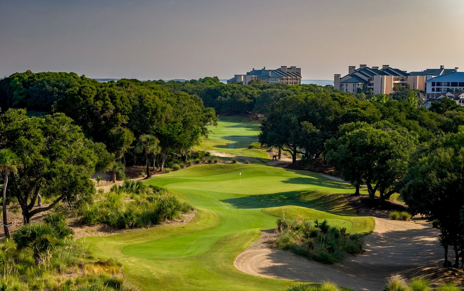 A lush golf course bites into a valley with trimmed fairways, sand bunkers, and towering trees, set against a distant row of buildings.