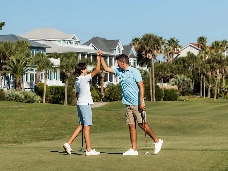 Two people on a golf course share a high-five after a game, with houses and palm trees in the background.