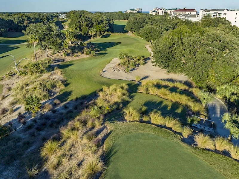 Aerial view of a golf course with greens, sand traps, and lush trees; some residential buildings are visible in the background, under a clear sky.