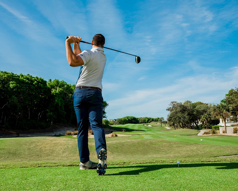 A golfer in a white shirt and blue jeans tees off on a sunny green course with trees lining the backdrop.