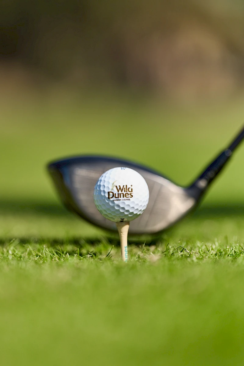 A golf ball on a tee with a club behind it on a grassy tee box, branded “WGI Dunes,” shot in focus against a blurred background.