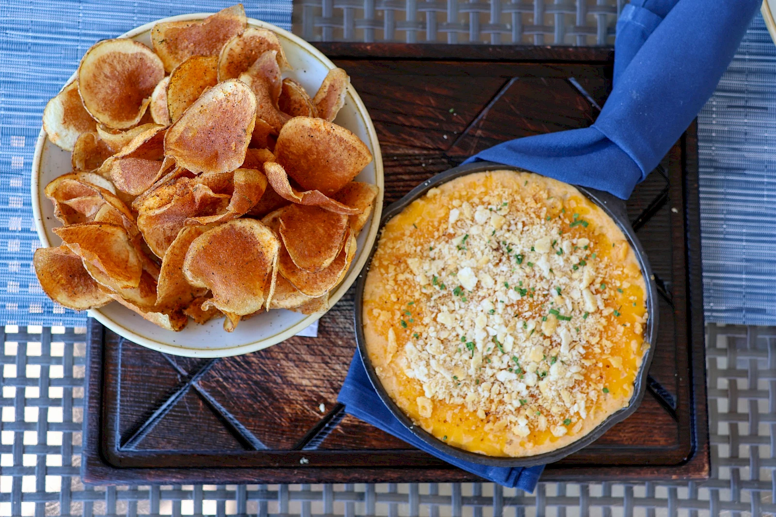 A bowl of potato chips and a small skillet with a cheesy herb dip on a outdoor tray, served with a blue napkin.