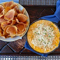 A bowl of potato chips and a small skillet with a cheesy herb dip on a outdoor tray, served with a blue napkin.