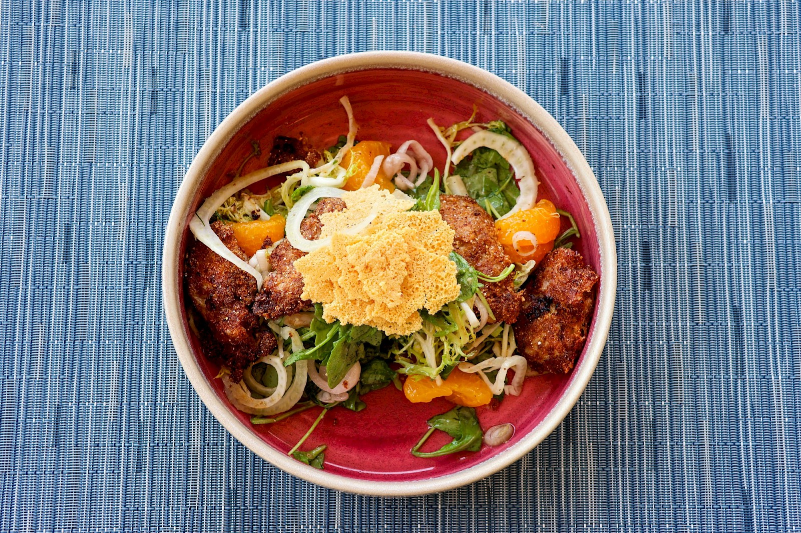 A colorful bowl of beef or meat with greens, onions, peppers, and a crispy topping on top, all on a blue placemat.
