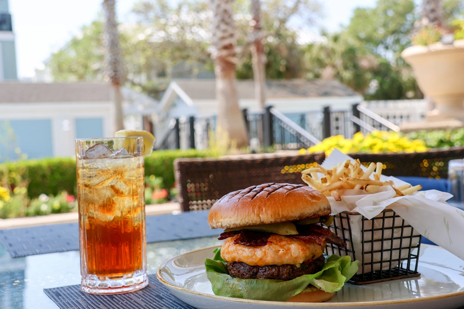 A burger with lettuce, a side of fries, and a glass of iced tea on an outdoor table, sunny day.