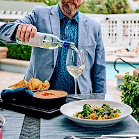 A man in a blue blazer pours white wine for two plates of food—one sizzling skillet, one pasta dish—on a sunny outdoor patio.