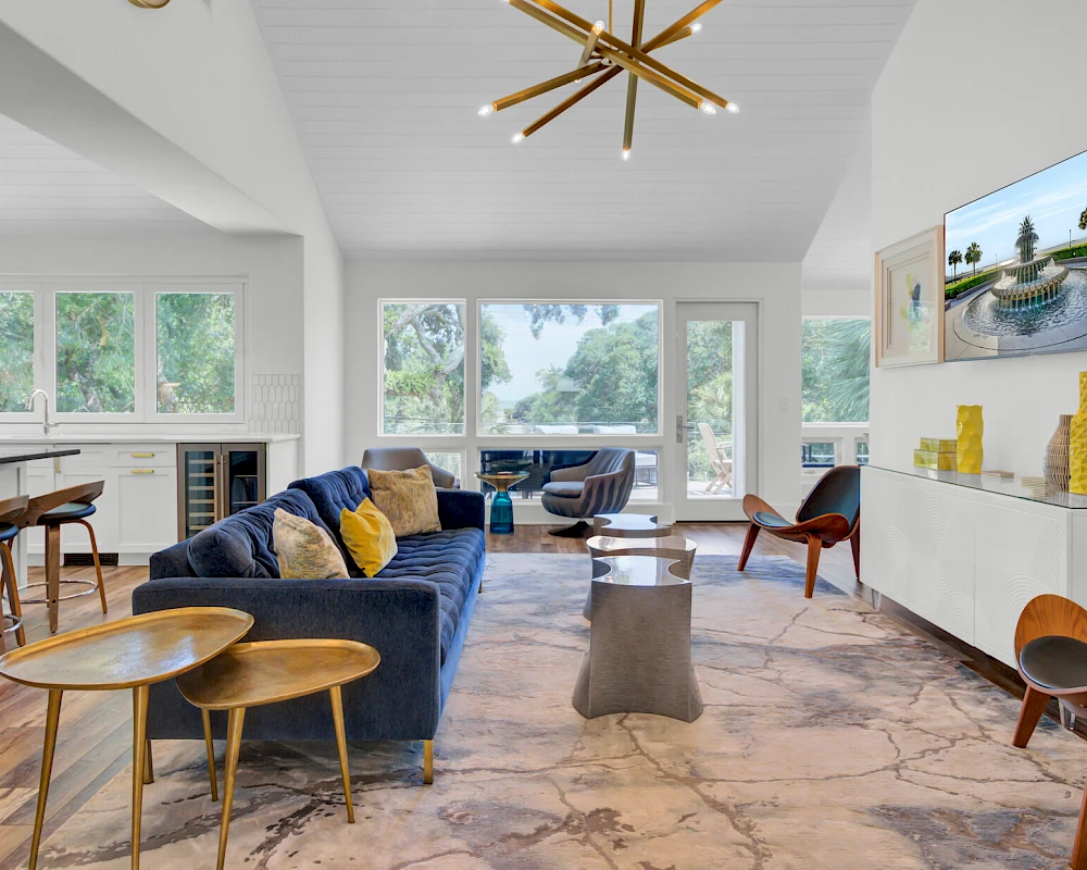 A modern, airy living room with a blue sofa, wooden side tables, marble floor, and a wall-mounted TV; large windows bring in natural light.