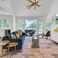 A modern, airy living room with a blue sofa, wooden side tables, marble floor, and a wall-mounted TV; large windows bring in natural light.