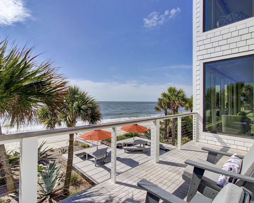 A sunny beachfront balcony with white railing, palm trees, and lounge chairs facing the ocean; a modern brick house windowed wall to the right.