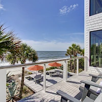 A sunny beachfront balcony with white railing, palm trees, and lounge chairs facing the ocean; a modern brick house windowed wall to the right.