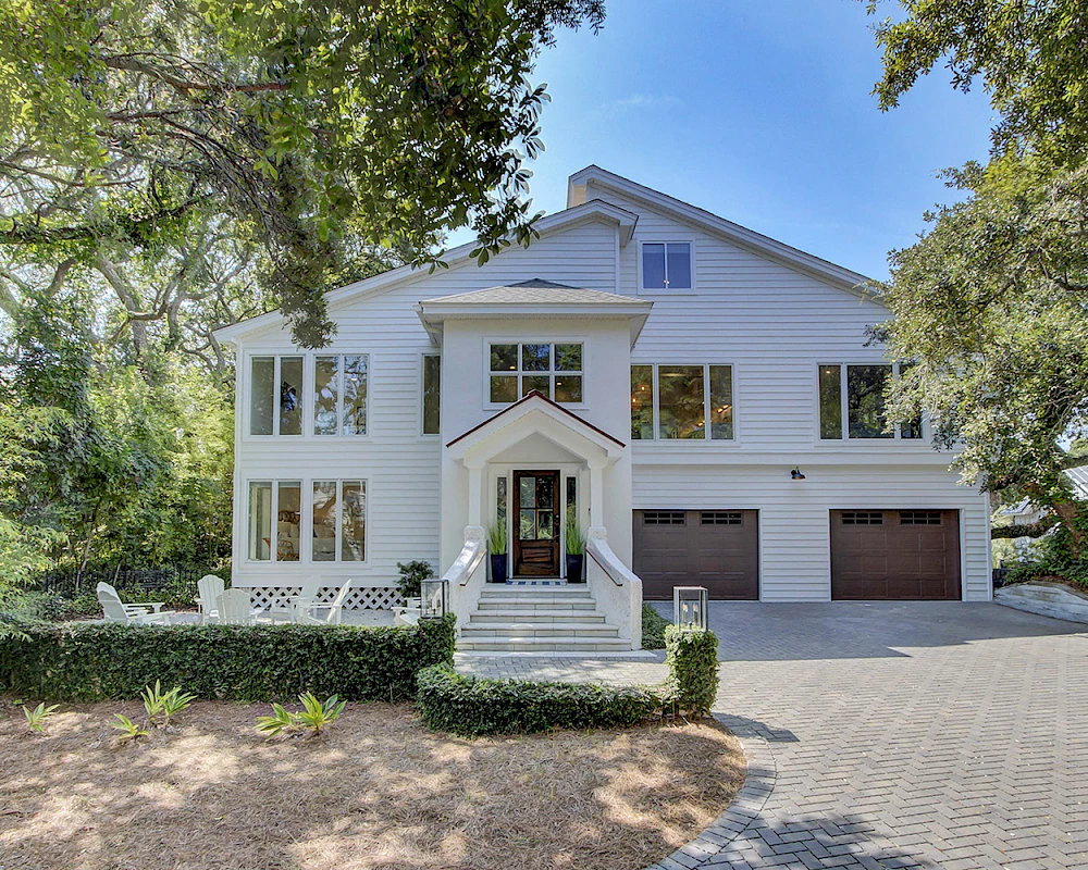 A large white two-story house with a peaked entry, multiple windows, a front porch, and a three-car garage, surrounded by trees and a paved driveway.