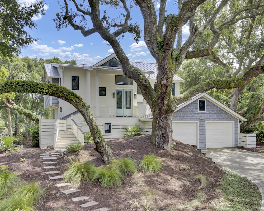 A two-story white house with a detached two-car garage, surrounded by mature trees and a landscaped yard, stone steps leading up the slope.