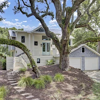 A two-story white house with a detached two-car garage, surrounded by mature trees and a landscaped yard, stone steps leading up the slope.