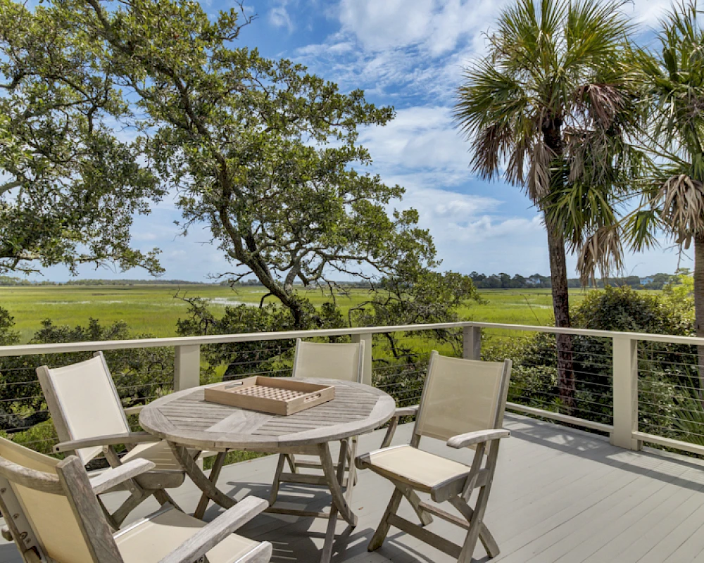 A wooden deck with a round table and six chairs overlooks a sunny landscape with trees and palm trees under a blue sky.