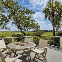 A wooden deck with a round table and six chairs overlooks a sunny landscape with trees and palm trees under a blue sky.