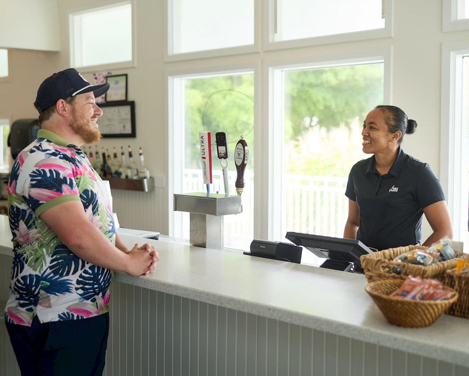 A man in a colorful floral shirt stands at a cafe counter chatting with a smiling cashier while a basket of snacks sits nearby.
