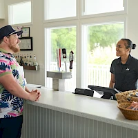 A man in a colorful floral shirt stands at a cafe counter chatting with a smiling cashier while a basket of snacks sits nearby.