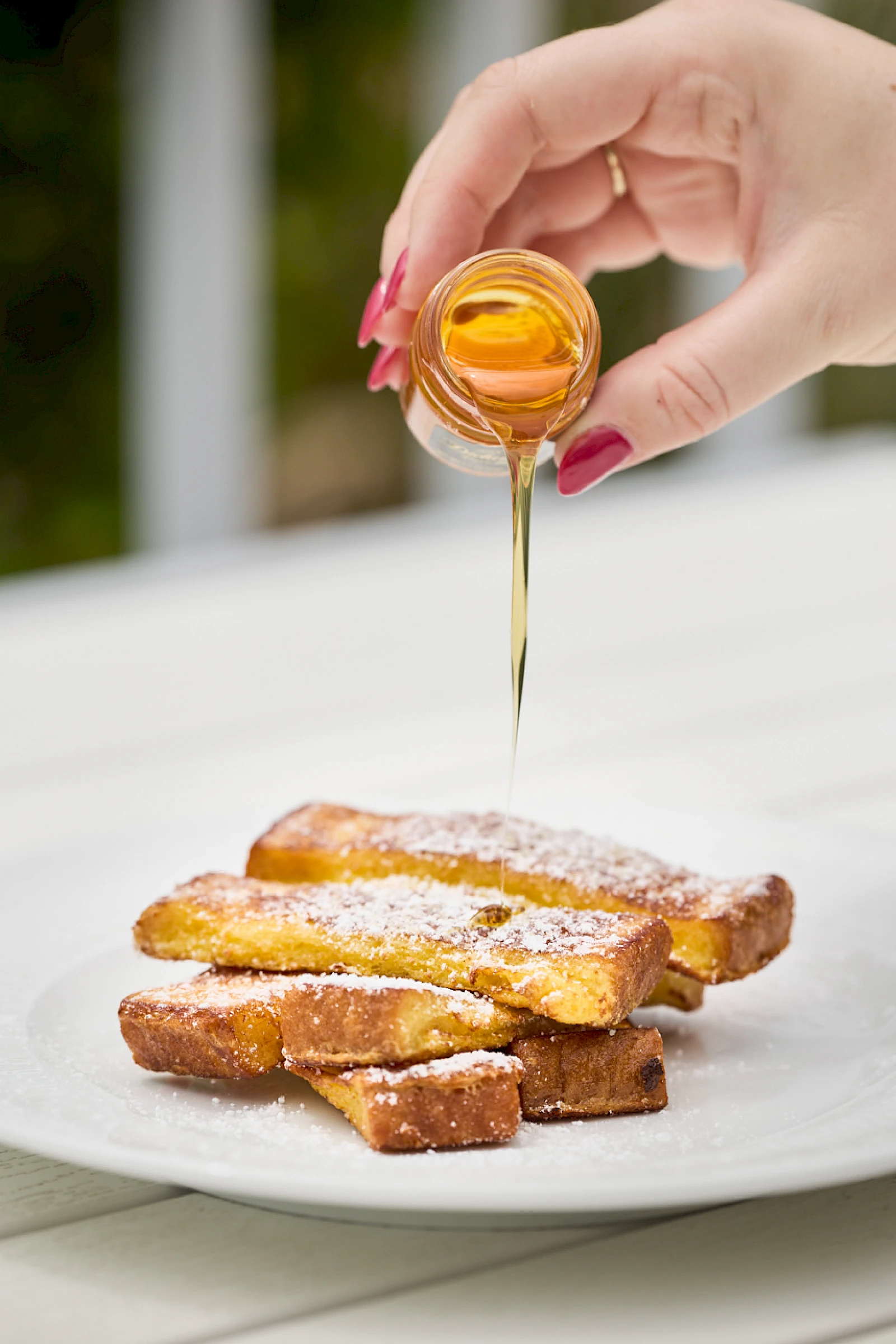 Pouring honey over a stack of French toast sticks on a white plate, drizzle cascading down with a sweet glaze ending deliciously.