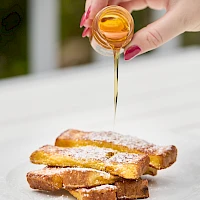 Pouring honey over a stack of French toast sticks on a white plate, drizzle cascading down with a sweet glaze ending deliciously.