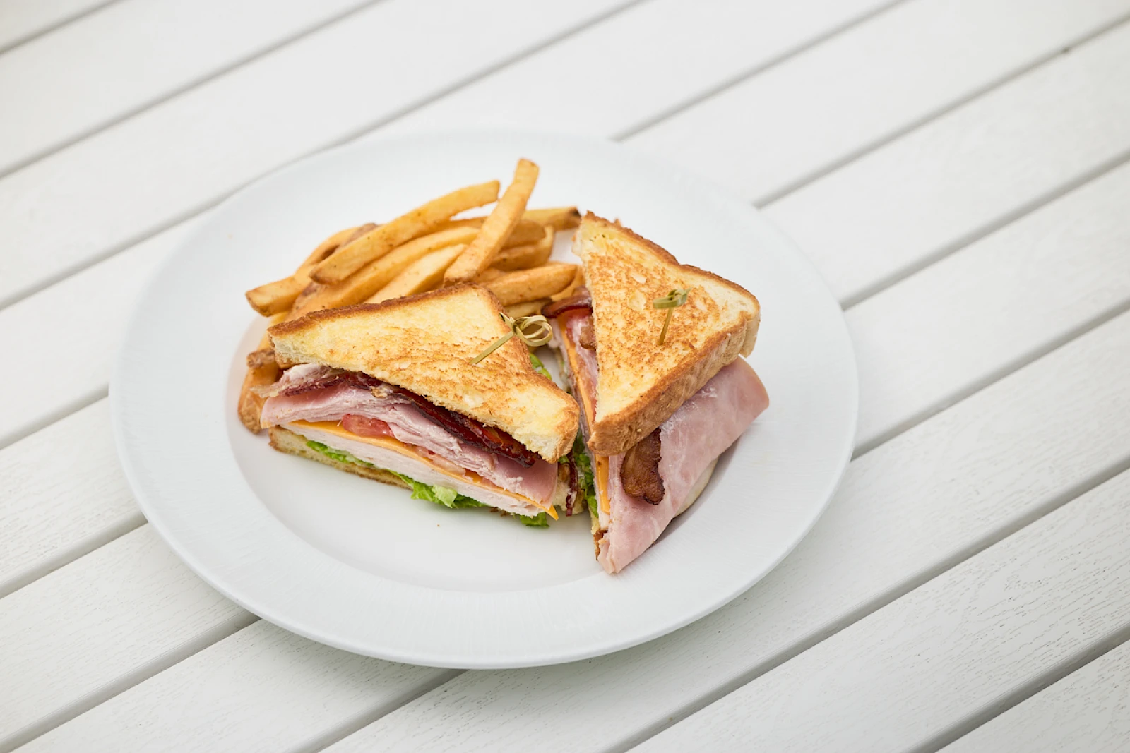 A plated ham and cheese club sandwich with crispy fries on the side, cut in half to reveal ham, cheese, lettuce, and tomato.