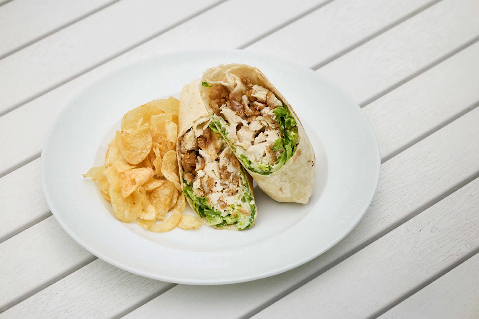 A plate with a wrap sandwich and a side of potato chips on a white table.