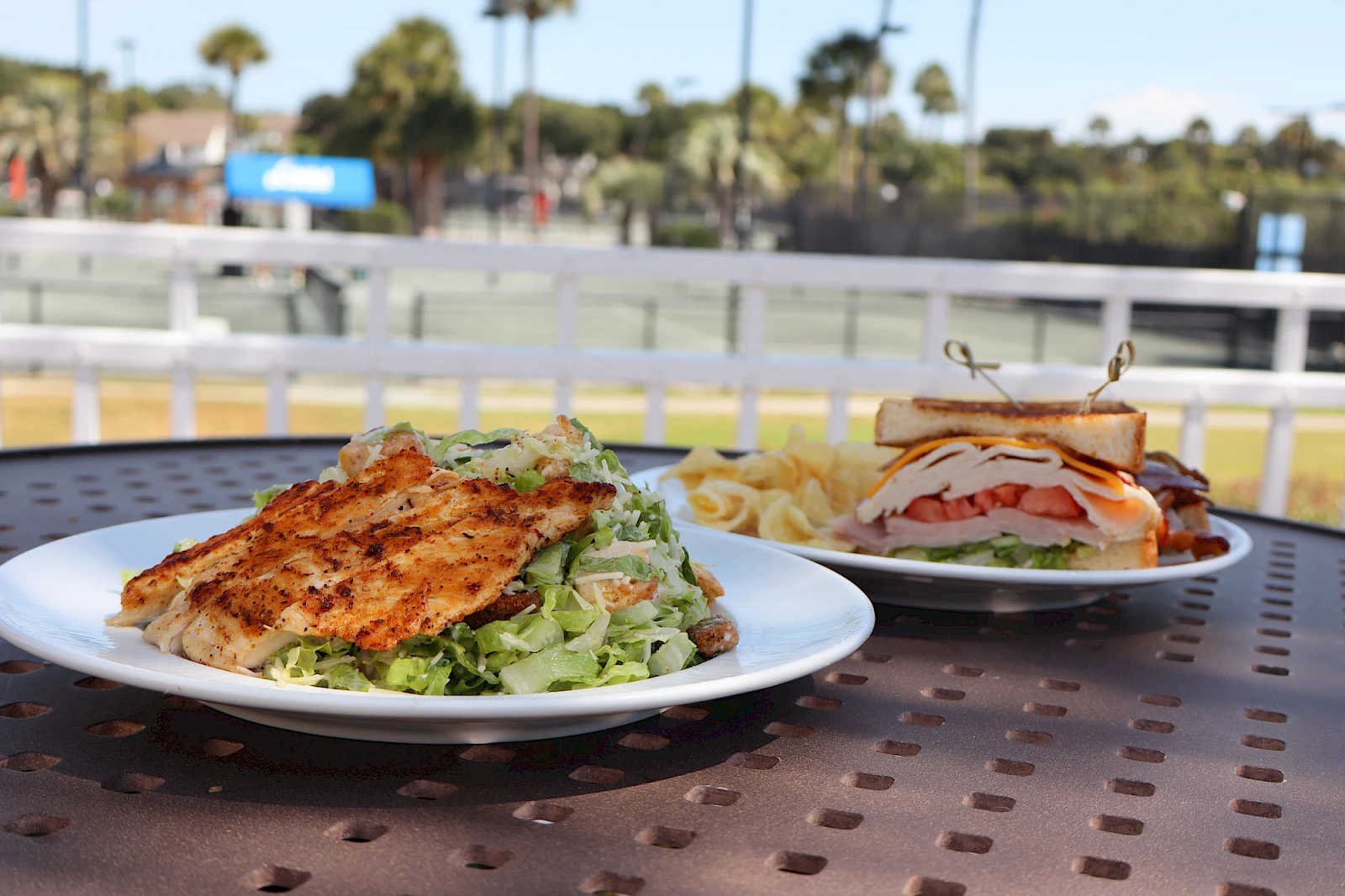 Two plates of food on a metal table: a grilled chicken sandwich with lettuce and a side salad on the left, and a club sandwich with tomatoes and bacon on the right, with fries in the background, outdoors at a sunny location.