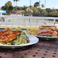 Two plates of food on a metal table: a grilled chicken sandwich with lettuce and a side salad on the left, and a club sandwich with tomatoes and bacon on the right, with fries in the background, outdoors at a sunny location.