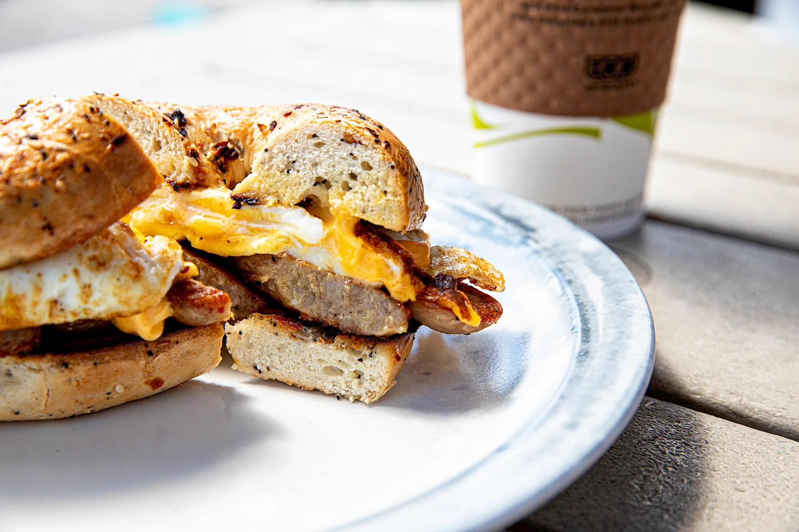 A breakfast sandwich with eggs, cheese, and bacon on toasted English muffins, beside a coffee cup on a wooden table.