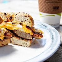A breakfast sandwich with eggs, cheese, and bacon on toasted English muffins, beside a coffee cup on a wooden table.