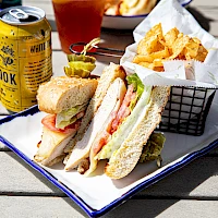 A plate with a club sandwich, a basket of chips, plus a can of White Claw and a drink on a wooden table.