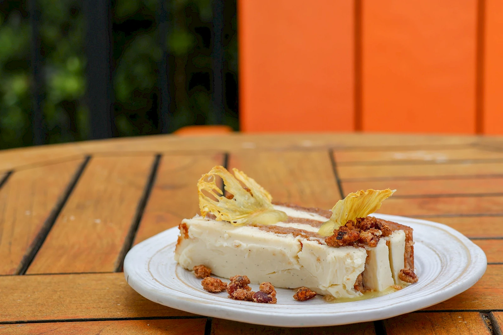 A slice of creamy cheesecake on a plate with pecans and decorative pineapple slices, set on a wooden table outdoors.