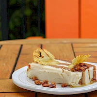 A slice of creamy cheesecake on a plate with pecans and decorative pineapple slices, set on a wooden table outdoors.