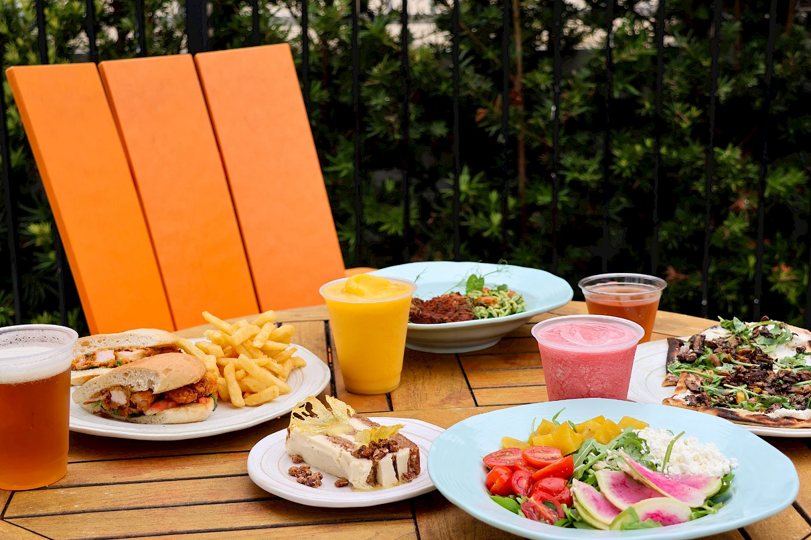 A sunny patio meal with burgers, fries, salad, dessert, a smoothie, and colorful drinks on a wooden table.