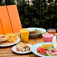 A sunny patio meal with burgers, fries, salad, dessert, a smoothie, and colorful drinks on a wooden table.