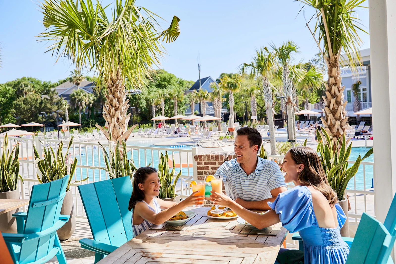 A family enjoys a sunny poolside meal at a resort, with palm trees, blue chairs, and a refreshing outdoor dining setup, smiling and sharing food.