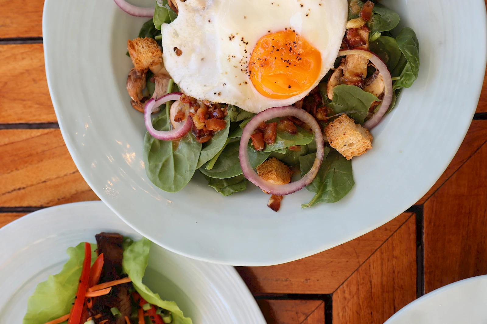 A fresh salad with baby greens, onions, croutons, bacon bits, and a sunny-side-up egg on top, with a light peppery finish.