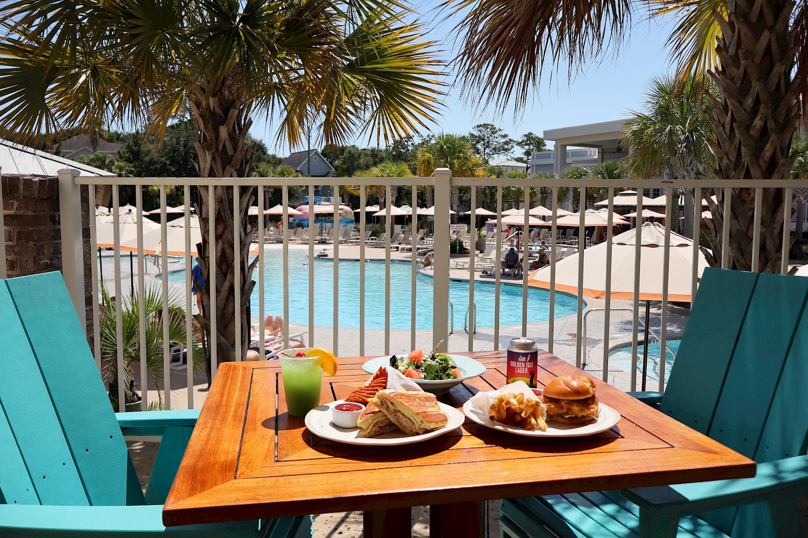 A sunny poolside breakfast scene with a wooden table, two plates of croissants and food, drinks, teal chairs, and palm trees surrounding a resort pool.