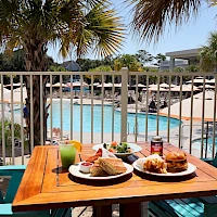 A sunny poolside breakfast scene with a wooden table, two plates of croissants and food, drinks, teal chairs, and palm trees surrounding a resort pool.