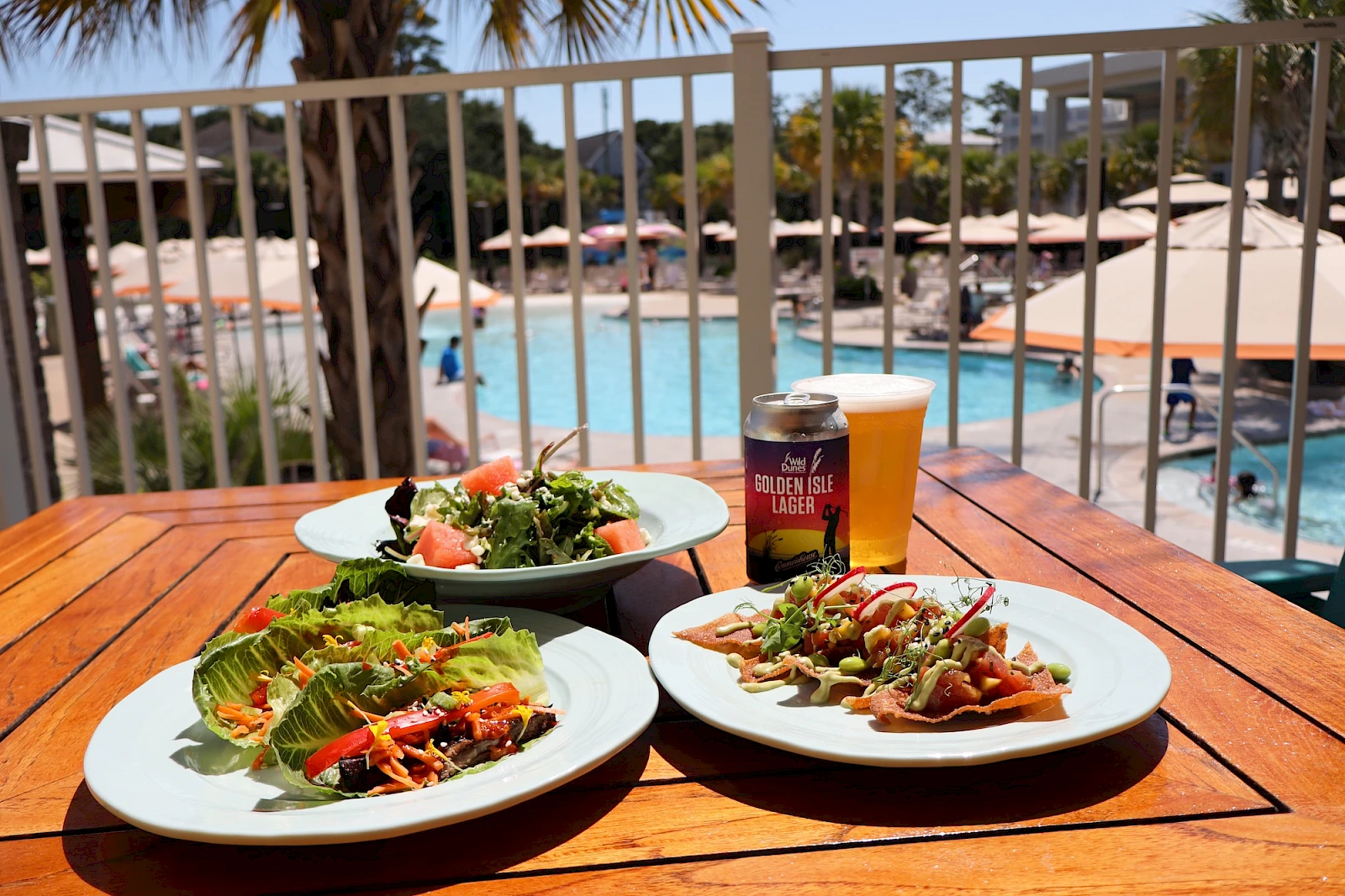 A poolside scene with two plates of colorful salad, a bowl of greens, and a beer on a wooden table near sunlit lounge chairs and umbrellas by the pool.