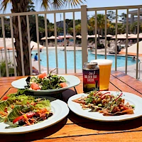 A poolside scene with two plates of colorful salad, a bowl of greens, and a beer on a wooden table near sunlit lounge chairs and umbrellas by the pool.