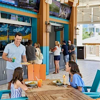 A sunny outdoor bar/restaurant with people casually dining and ordering drinks at a bright, tropical-looking venue.