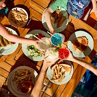 A table full of friends sharing a colorful picnic: bowls of fries, veggies, dips, and bright drinks as they dip and toast together.
