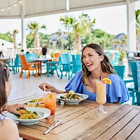 Two women dine at a sunny outdoor restaurant, sharing smiles and drinks; colorful chairs surround wooden tables under string lights.