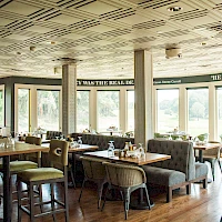 A bright, airy restaurant or cafe with large windows, wooden floors, and a mix of cushioned booths and chairs; daytime dining setup.