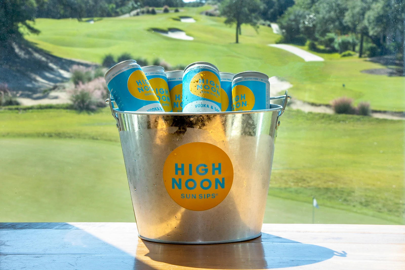 A metal bucket filled with High Noon hard seltzer cans sits on a table outdoors, a sunny golf course background behind.