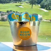 A metal bucket filled with High Noon hard seltzer cans sits on a table outdoors, a sunny golf course background behind.