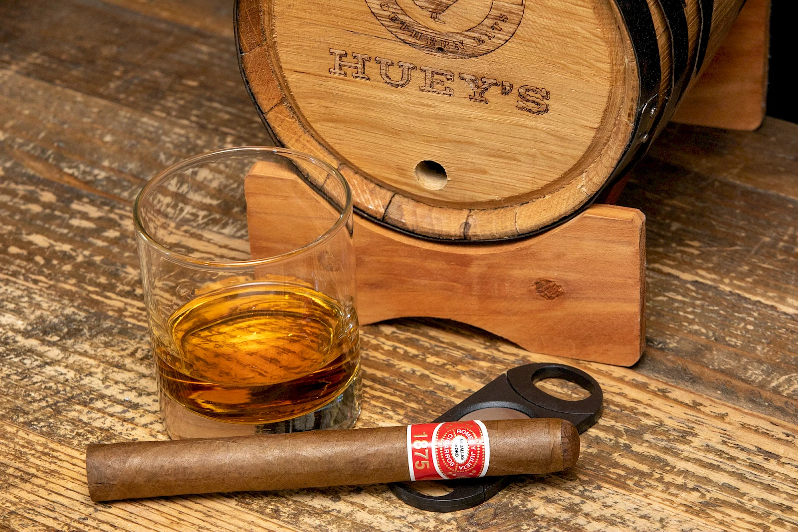 A whisky barrel, a glass with whiskey, a smoking cigar, and a cigar cutter resting on a weathered wooden table.