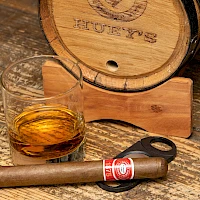 A whisky barrel, a glass with whiskey, a smoking cigar, and a cigar cutter resting on a weathered wooden table.